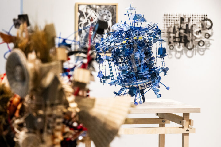 A blue abstract sculpture made of various small objects sits on a wooden table in an art gallery, with other mixed-media artworks and framed pieces displayed in the background of the Mills College Art Museum.