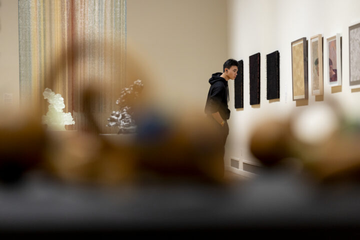 A young man in a black hoodie views framed artwork at the Mills College Art Museum, with sculptures and other visitors blurred in the foreground.