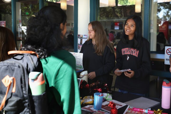Two students staffing a table at an outdoor campus event at Rothwell Center on Northeastern's Oakland campus talk with a student in a green sweatshirt