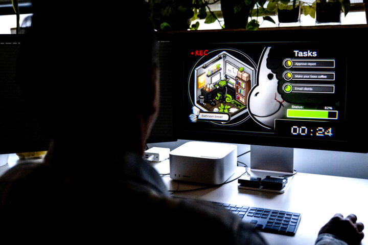A person seen from behind, in silhouette, plays a video game on a desktop monitor. The game shows a green chameleon working in an isometric office, with a task list and a countdown timer.