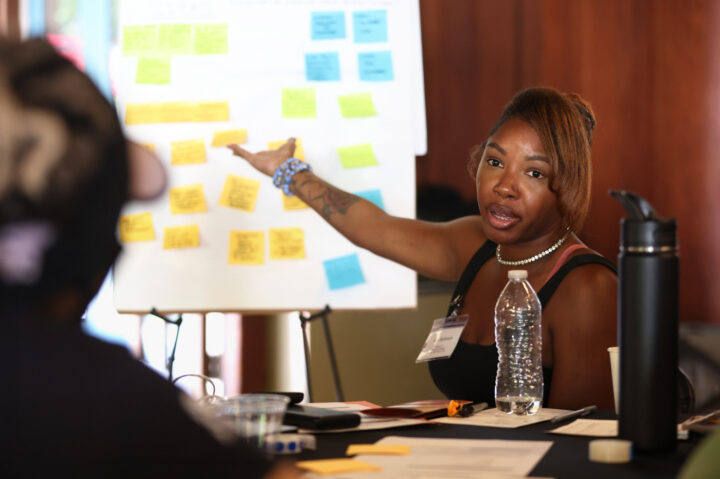 A woman gestures toward a presentation board covered with colorful sticky notes during a group workshop or meeting.