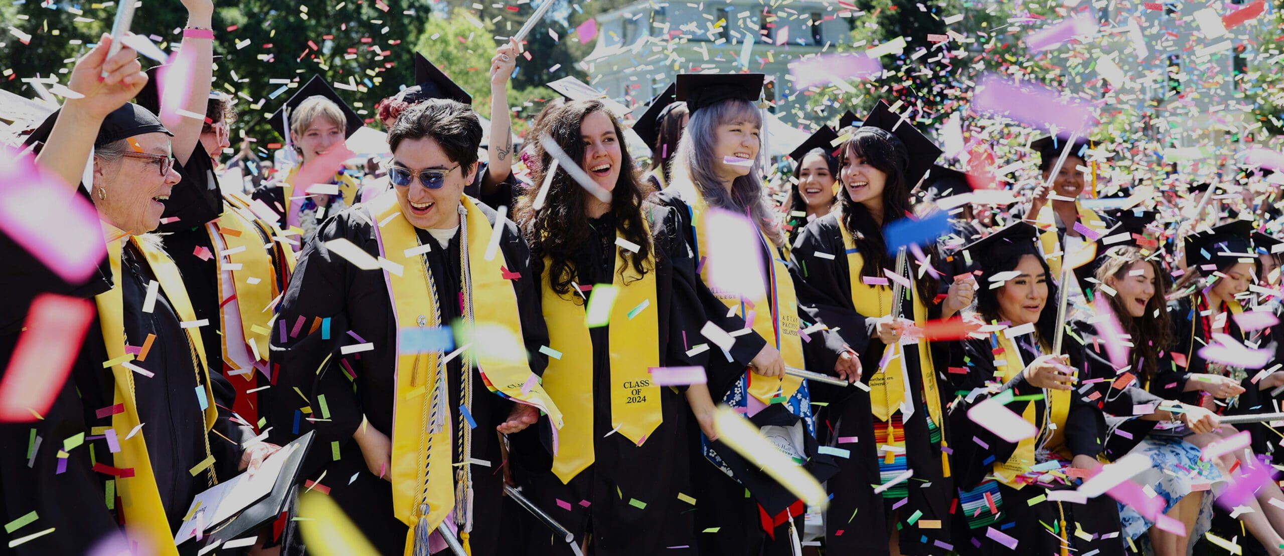 A group of graduates in caps and gowns celebrate outdoors, smiling and laughing as colorful confetti falls around them.