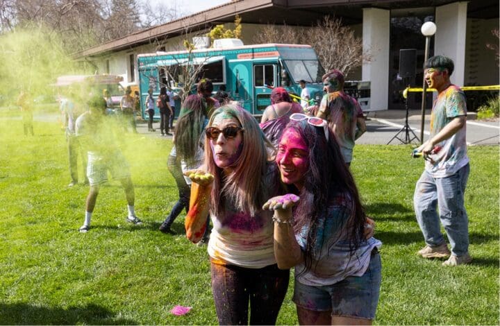 Two people stand close together on a grassy lawn, blowing colorful powder toward the camera during a Holi celebration. Others in the background, covered in powder, play and laugh near a food truck.