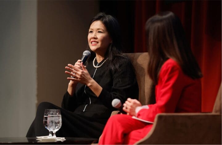 Two women sit on stage in conversation. The woman on the left, dressed in black, speaks into a microphone with a smile, while the woman on the right, dressed in red, listens attentively. A glass of water sits on the table nearby.