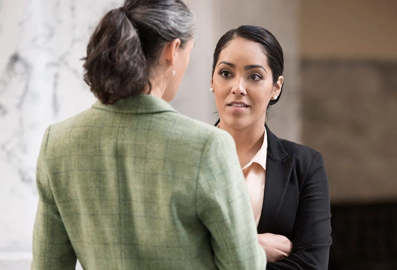 Two professionally dressed women are having a serious conversation indoors; one woman faces the camera with arms crossed, while the other faces away, showing only her back and side profile.
