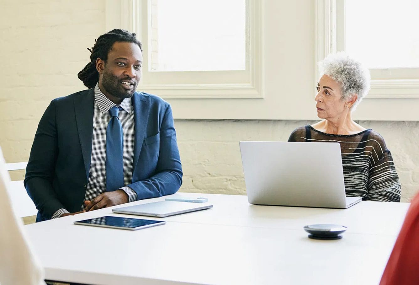 Two professionals sit at a conference table. A man in a suit speaks, while a woman with short gray hair and a striped shirt listens, her laptop open in front of her. Sunlight streams through nearby windows.