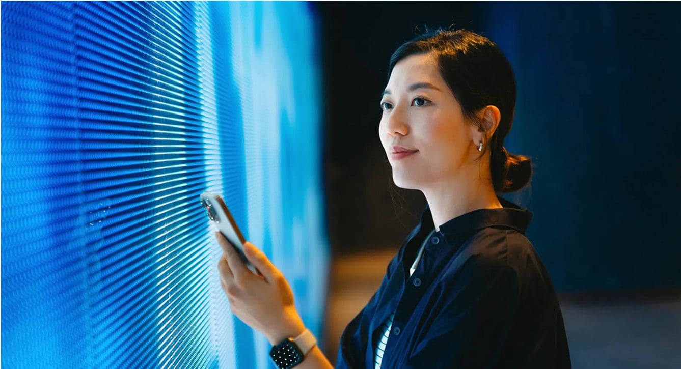 A woman in a dark shirt holds a smartphone and looks thoughtfully at a wall of blue LED lights in a modern, dimly lit space.