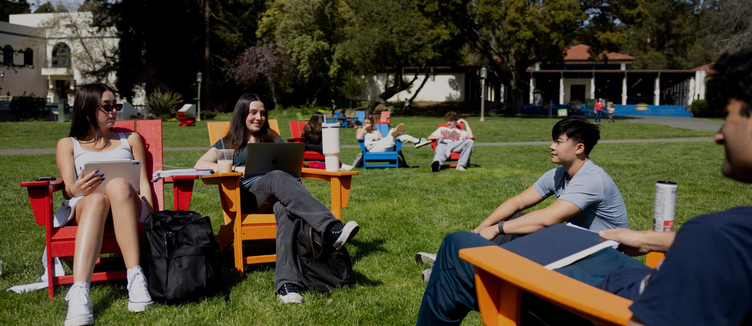 Four students sit on colorful Adirondack chairs in a grassy outdoor area, studying and talking together, with other people relaxing on chairs in the background and campus buildings nearby.