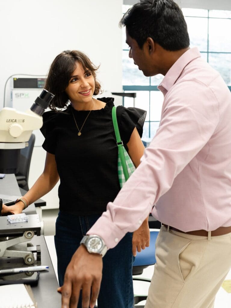 A woman and a man stand in a bright laboratory near a microscope. The woman, smiling, wears a black top and carries a green bag. The man, in a light pink shirt, gestures while speaking to her.