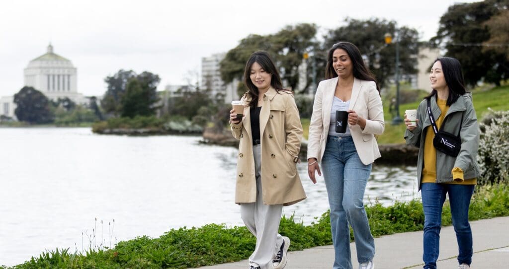 Three women walk along a lakeside path, smiling and holding coffee cups, with trees and a domed building in the background on a cloudy day.