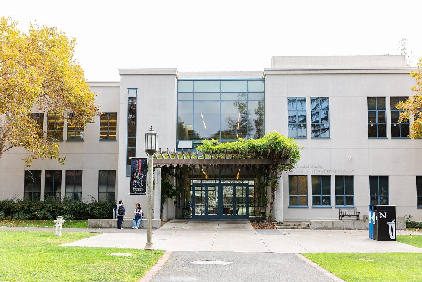 The Betty Irene Moore Natural Sciences building with large windows, a vine-covered pergola above the entrance, and a few people standing outside. Trees with autumn leaves line the walkway leading to the door.