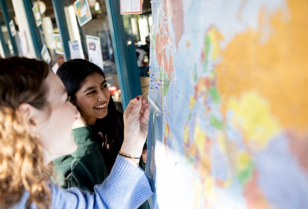 Two young women smile while pinning locations on a large world map, with one woman placing a pin and the other looking on. The scene is bright and appears to take place in a classroom or community space.