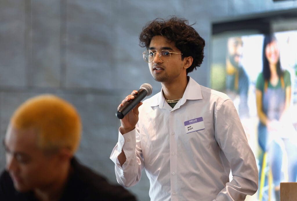 A young man with glasses and a white shirt holds a microphone and speaks at an indoor event. He wears a name tag. Another person with blond hair is blurred in the foreground.