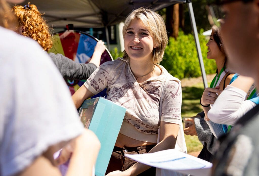 A smiling young woman holds a large turquoise box at an outdoor event, surrounded by other people and a colorful wheel under a canopy, with sunlight and greenery in the background.