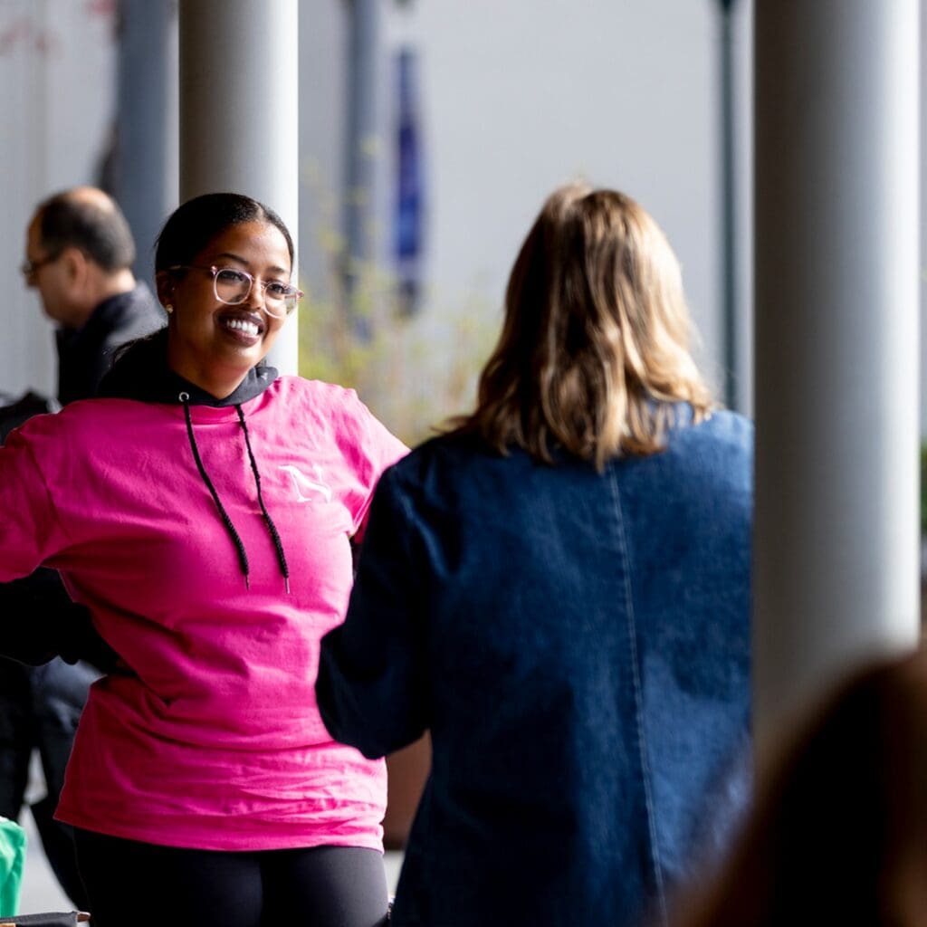 A woman in a bright pink hoodie smiles while talking to another person wearing a blue jacket. They are standing outside, and other people are visible in the background.