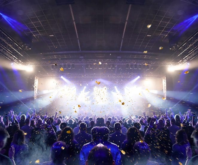A large crowd enjoys a concert in an indoor venue, with bright stage lights shining and confetti falling from above, creating an energetic and festive atmosphere.