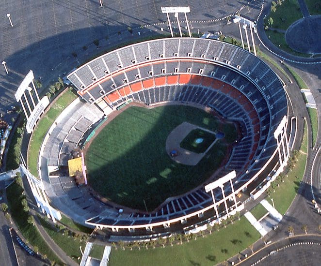Aerial view of an empty baseball stadium with green field, brown infield, and surrounding seating areas. Some seats are shaded, and the parking lot around the stadium appears mostly empty.