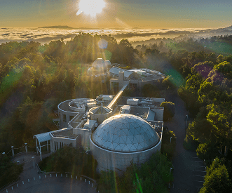 Aerial view of a modern building with a glass dome roof, surrounded by trees and greenery, with sunlight streaming through morning mist and clouds in the background.