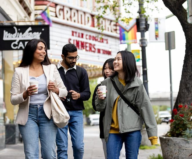 Four young adults walk together on a city sidewalk, smiling and holding coffee cups. A theater marquee and rainbow flags are visible in the background, creating a lively urban atmosphere.