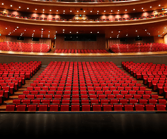 A view from the stage of an empty theater auditorium, featuring rows of red seats and multiple balconies with warm lighting.