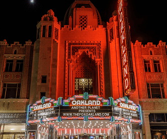 Night view of the historic Fox Theater in Oakland, California, illuminated with red lights and a bright marquee displaying upcoming performances. The building features ornate architectural details and a large vertical sign reading "Oakland.