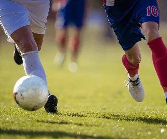 Close-up of two soccer players running towards a white soccer ball on a grassy field, with their legs in motion and other players blurred in the background.