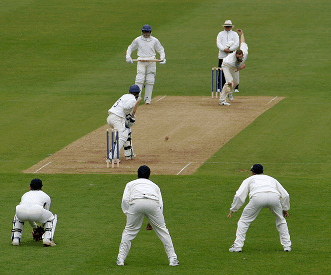 A cricket bowler delivers the ball to a batsman, with the wicketkeeper and fielders positioned around the pitch, while an umpire observes the play. All players are dressed in traditional white uniforms.