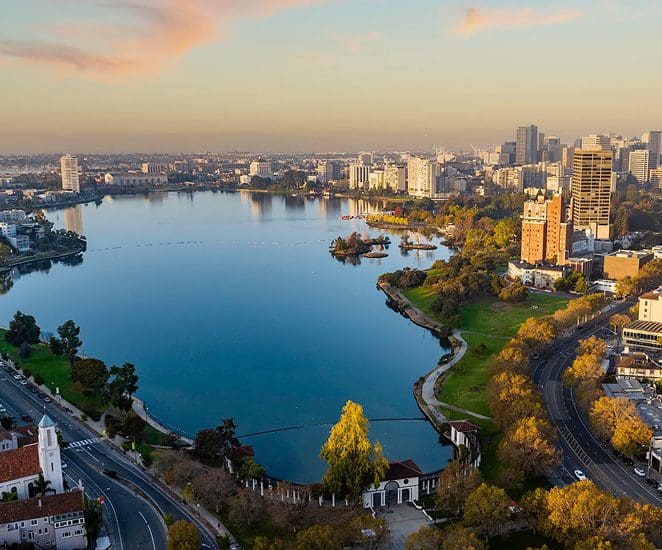 Aerial view of a large urban lake surrounded by trees, parks, and city buildings at sunset, with clear skies and roads winding around the water.