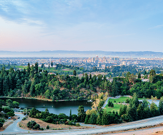 A scenic view overlooking a lush, green park with a small lake, surrounded by trees. In the distance, a city skyline rises under a clear sky, with mountains visible on the horizon.