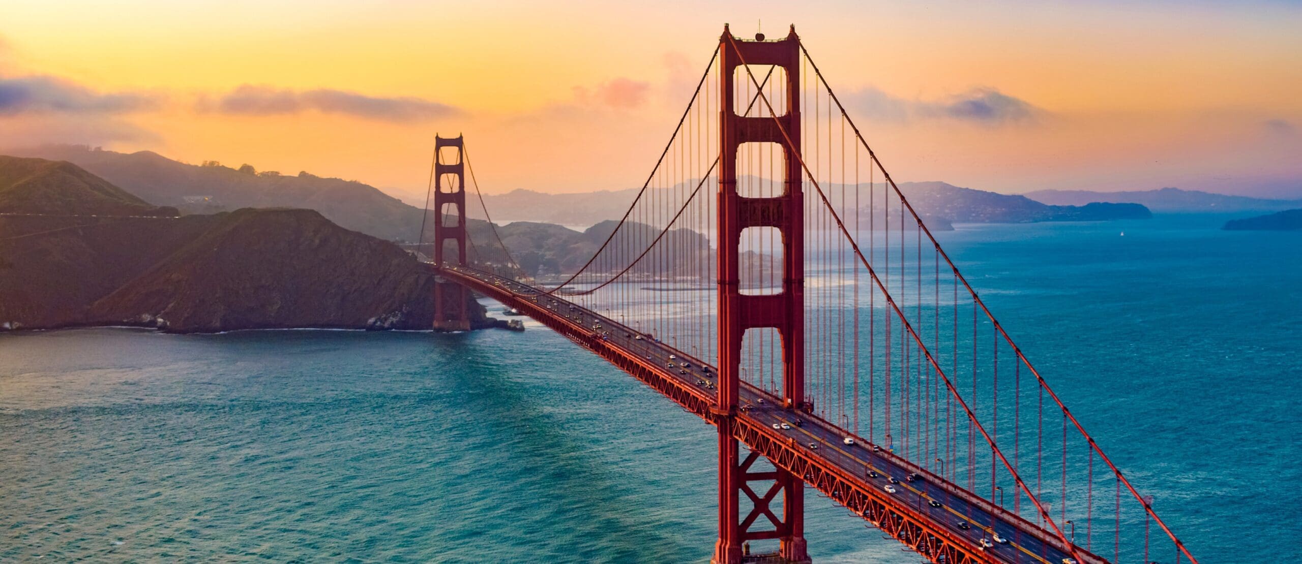 The Golden Gate Bridge stretches over blue water at sunset, with orange towers and cables visible. Hills and coastline are in the background under a colorful sky with clouds.
