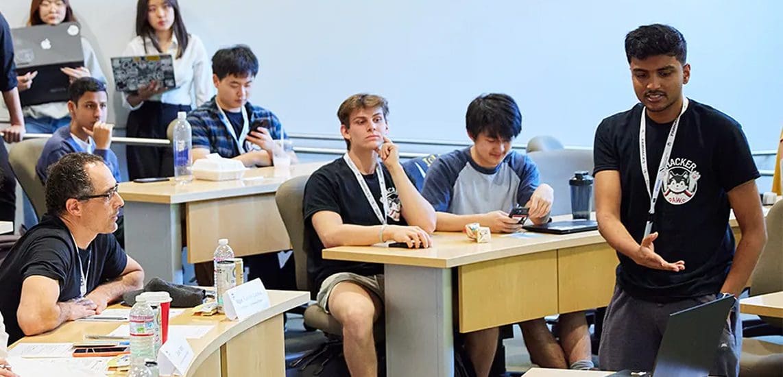 A young man stands and speaks in front of a classroom, while other students sit at desks listening. A teacher sits to the left, and several students have laptops and notebooks open.