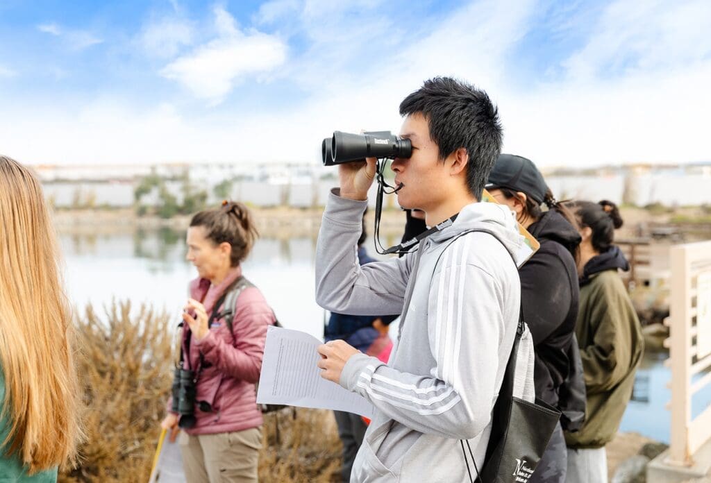 A young man holding binoculars and a paper watches birds by a lake with a group of people on a bright, partly cloudy day. Other group members also have binoculars and are looking in different directions.