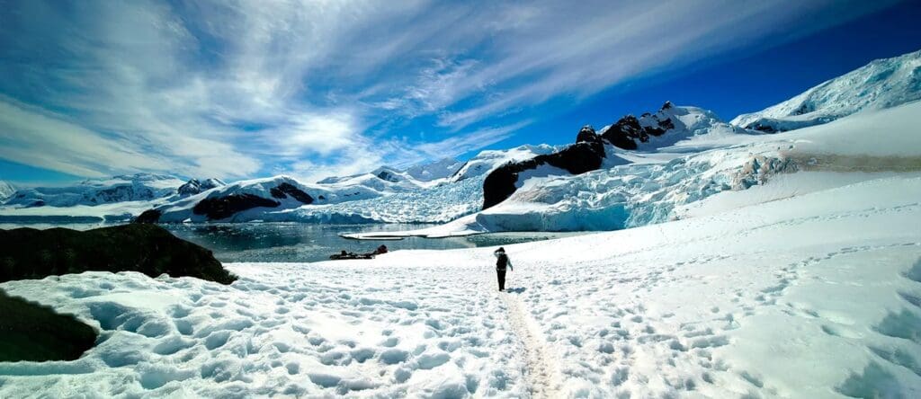 A person stands on a snowy landscape surrounded by icy mountains and a partly frozen lake under a blue sky with wispy clouds.