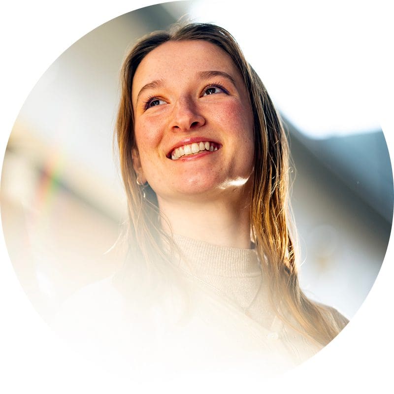 A young woman with long, light brown hair smiles and looks up, bathed in warm sunlight. She is wearing a light turtleneck sweater, and the background is softly blurred.