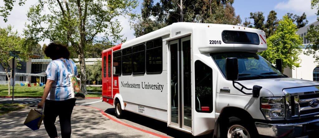 A person walks toward a white Northeastern University shuttle bus parked on a campus road surrounded by trees and greenery on a sunny day.