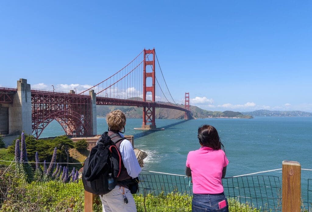 Two people stand near a fence, overlooking the Golden Gate Bridge in San Francisco on a sunny day, with blue sky, ocean, and hills in the background.