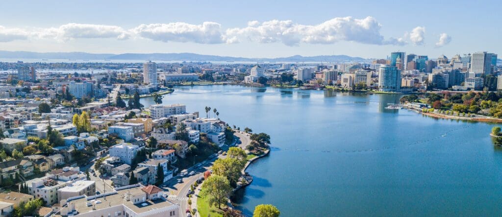 Aerial view of Lake Merritt surrounded by buildings and greenery in Oakland, California, with the city skyline and hills visible under a blue sky with scattered clouds.