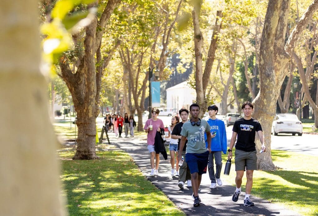 A group of students walks along a tree-lined sidewalk on a sunny day, carrying backpacks and water bottles, with parked cars and more people visible in the background.