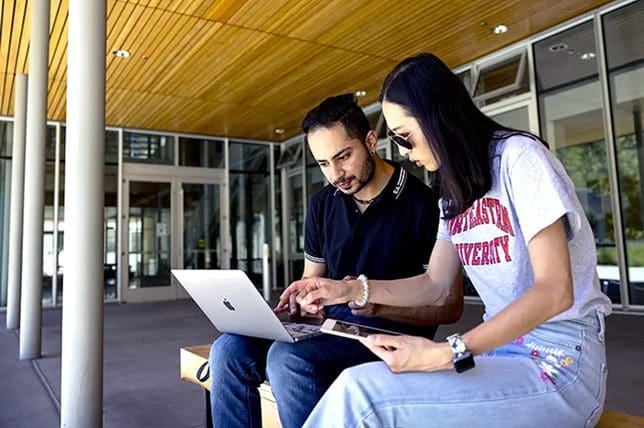 Two people sit on a bench outside a the Lorey I. Lokey Graduate School of Business building on Northeastern's Oakland campus, looking at a laptop together. One person points at the laptop screen while the other watches attentively. Both appear focused and engaged in their task.
