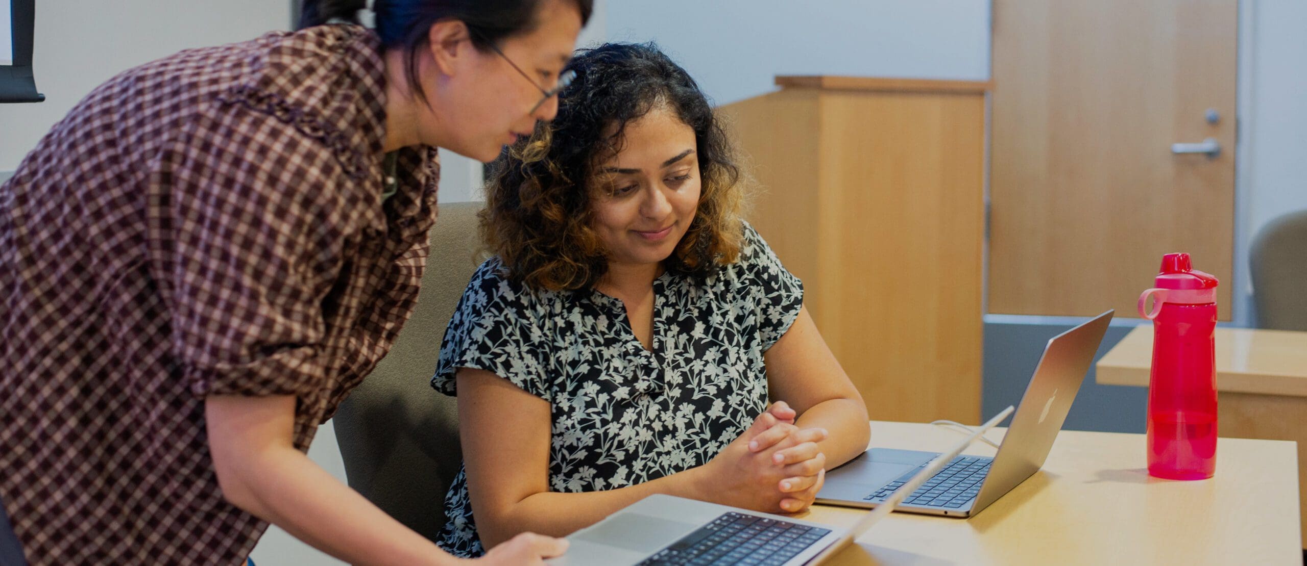 Two women work at a table with laptops. One stands and points at a laptop screen while the other sits, watching attentively. A red water bottle is on the table. The room has wooden furniture and a closed door.