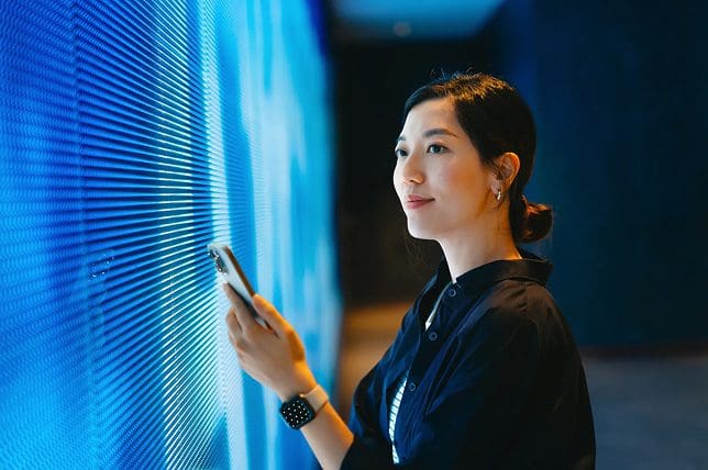 A woman holding a smartphone stands next to a glowing blue wall, looking ahead with a slight smile. She is wearing a dark button-up shirt and a smartwatch. The setting appears modern and futuristic.