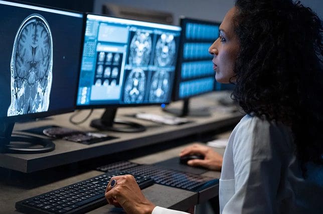 A woman in a lab coat analyzes brain scans displayed on multiple monitors at a workstation, using a mouse and keyboard in a dimly lit room.