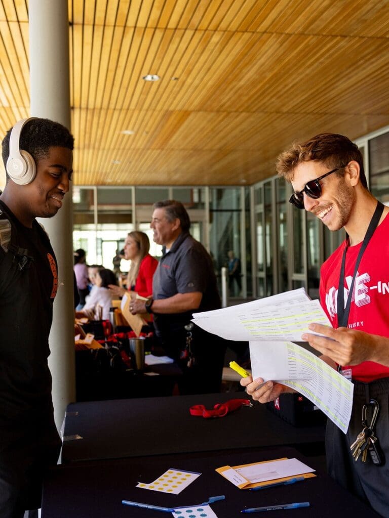 A smiling man in sunglasses and a red T-shirt holds papers and a highlighter while talking to a young man wearing headphones at a check-in table indoors. Other people are visible in the background.