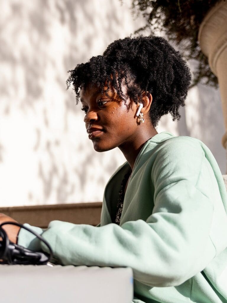 A young woman with curly hair and AirPods, wearing a light green sweater, sits outdoors in sunlight, looking focused while working on a laptop. Shadows of leaves are visible on the wall behind her.