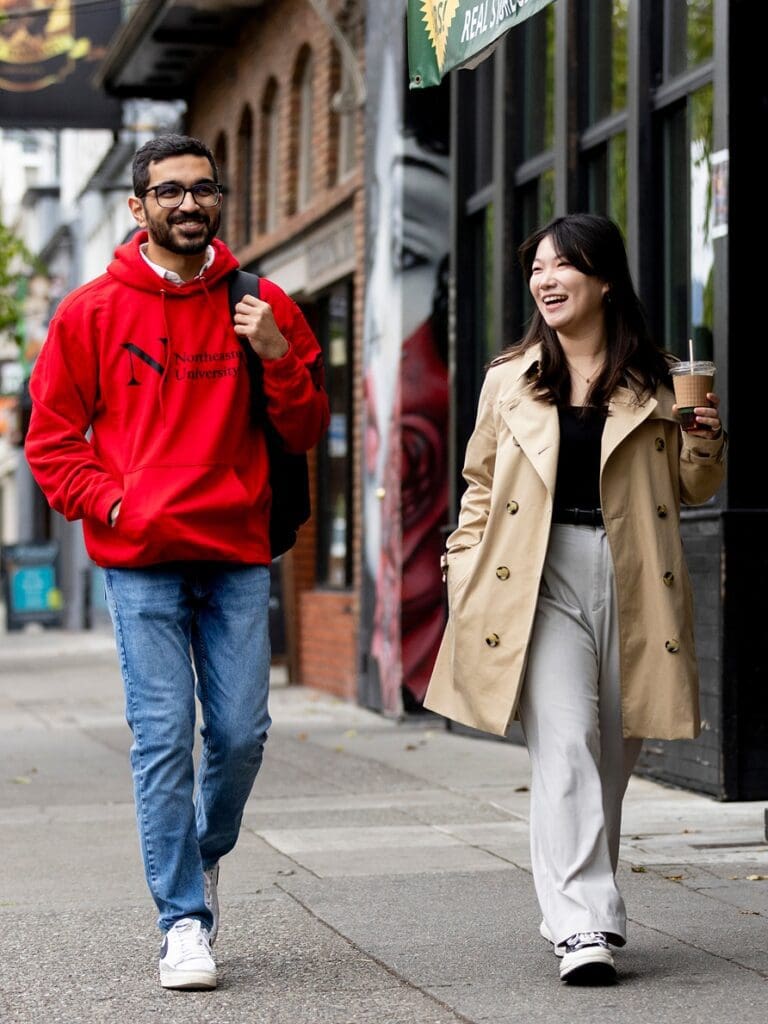 A man in a red hoodie and jeans and a woman in a beige trench coat and white pants walk side by side on a city sidewalk, smiling and talking. The woman holds an iced drink. Urban buildings line the street.