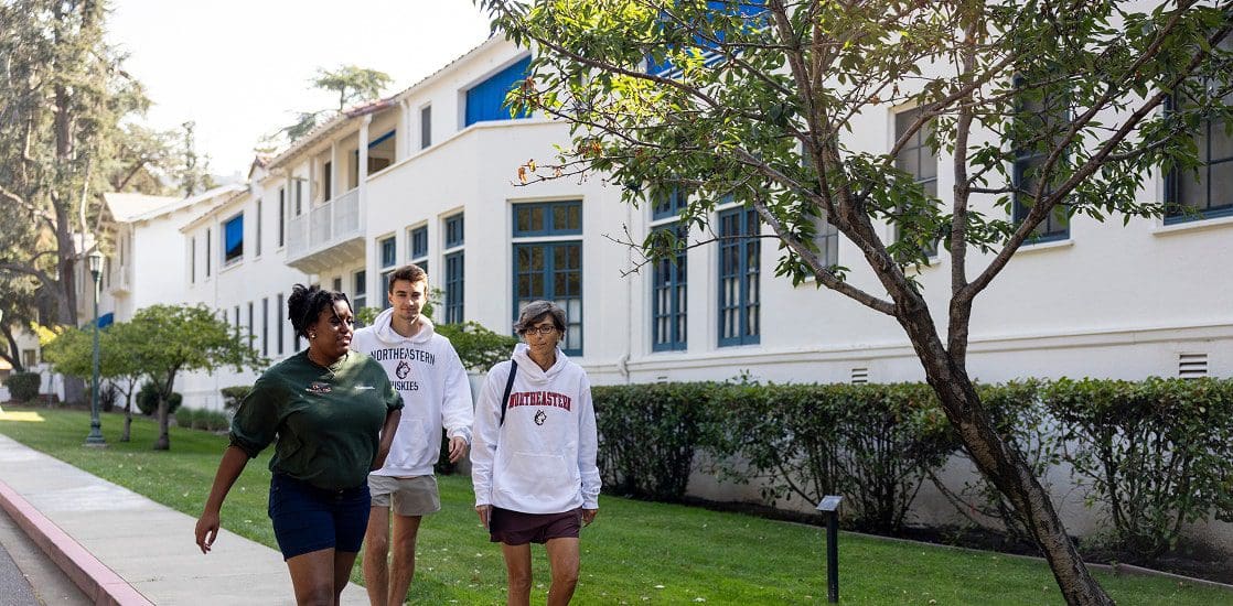 Three students walk together on a sidewalk beside a white building with blue window frames, surrounded by green bushes and trees. They appear to be talking and enjoying each other's company.