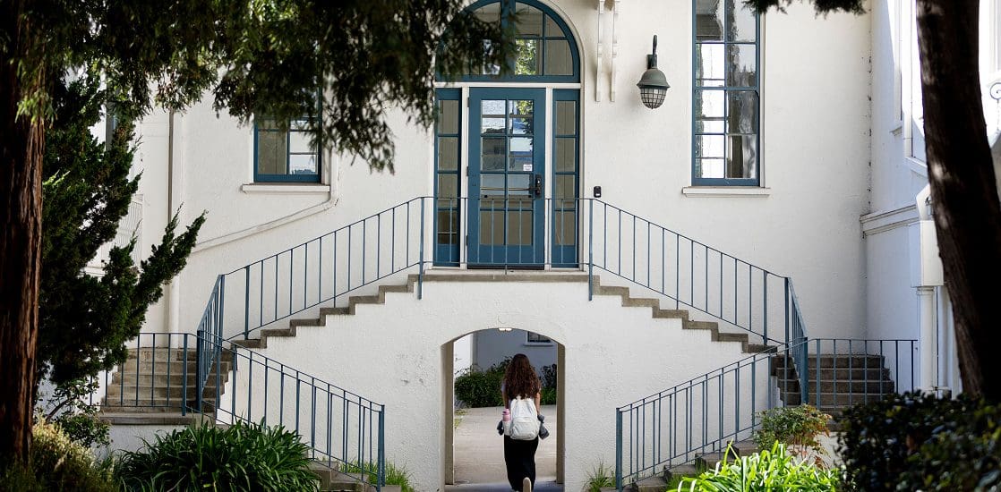 A person with a backpack walks toward a white building with blue double doors, symmetrical staircases, and arched entryways, surrounded by greenery and shaded by trees.