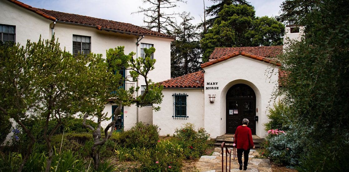 A person in a red jacket walks toward the entrance of a white stucco building with a red tiled roof named "Mary Morse," surrounded by lush greenery and trees.