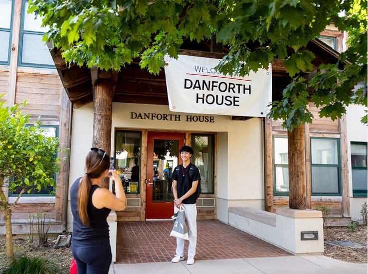 A woman takes a photo of a smiling young man standing under a "Welcome to Danforth House" banner in front of a building entrance shaded by green tree leaves.