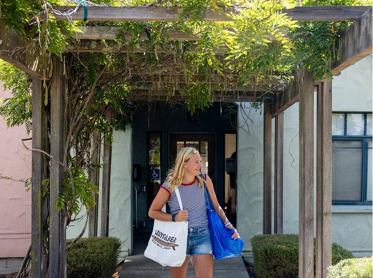 A young woman with blonde hair, wearing a striped shirt and denim shorts, walks under a wooden pergola with vines, carrying two bags and smiling outside a building.